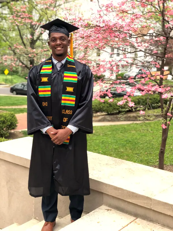 A young man in a graduation gown and stole in front of a flowering tree.